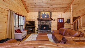 A log cabin interior with a stone fireplace, leather sofas, and a television. - Holiday Road! Cabin with Beautiful Outdoors, Game Room and Hot Tub (Broken Bow)
