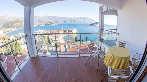 A balcony with a view of a coastal town, a table with a yellow tablecloth, and metal chairs. - Lux Panoramic Apartment (Budva)