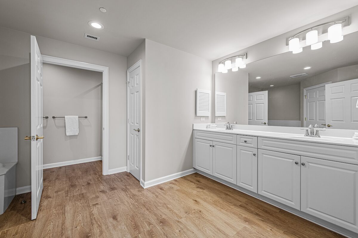 A bathroom with white cabinetry, a large mirror with lights, and a wooden floor.