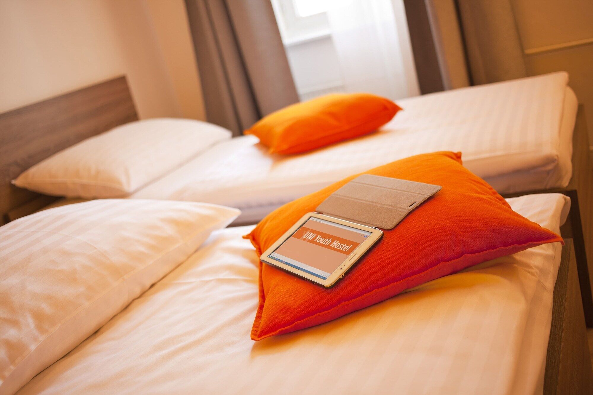 Zwei Betten mit weißen Laken und orangefarbenen Kissen, eines mit einem Tablet, das 'VINI Youth Hostel' anzeigt.