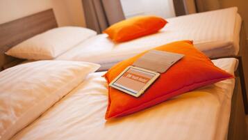 Zwei Betten mit weißen Laken und orangefarbenen Kissen, eines mit einem Tablet, das 'VINI Youth Hostel' anzeigt.
