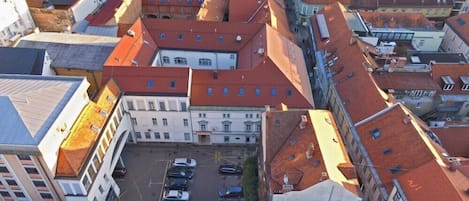 An aerial view of a cityscape with a prominent building featuring a red-tiled roof, a courtyard with parked cars, and adjacent buildings with similar architectural styles.
