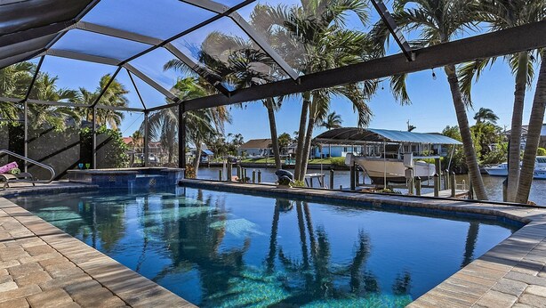 Piscina límpida com borda azulejada, rodeada por palmeiras e um espaço coberto.