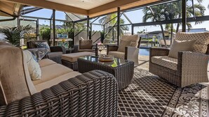 Área de estar interna iluminada pelo sol, com móveis de vime, mesa de centro de vidro e grandes janelas com vista para palmeiras e uma piscina.