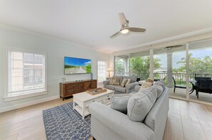A living room with a sofa, coffee table, and TV, a balcony with a table and chairs, and large windows.