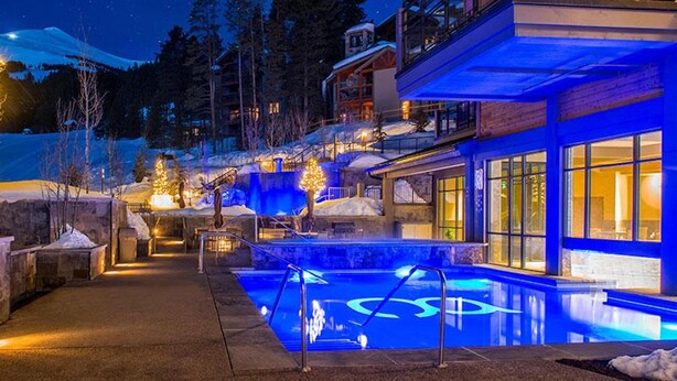A modern building with a pool area, illuminated by blue lights, surrounded by snow-covered landscape and trees.