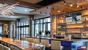 A modern bar area with a long wooden counter, bar stools, and a television screen displaying a sports event.