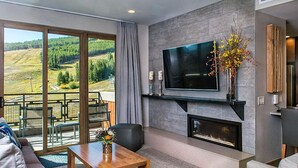 A modern living room with a large TV, fireplace, and a view of a green landscape through large windows.