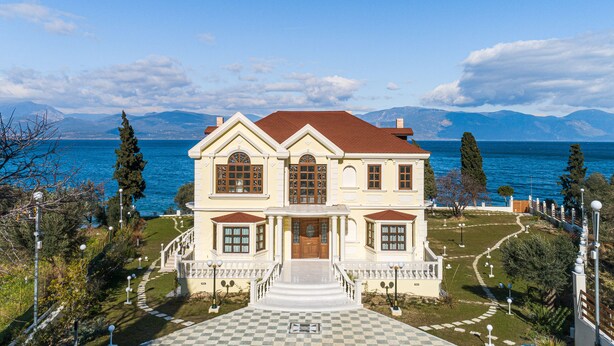 A large, two-story house with a checkered pathway leading to the entrance, situated by a body of water with mountains in the background. - Villa Filoxenia - by the sea - up to 12 guests (Aigialeia)
