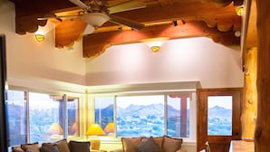 Ein gemütliches Wohnzimmer mit Holzbalkendecke, Deckenventilator, großem Fenster mit Bergblick, einer Couch, einem Couchtisch und einem Essbereich mit einem Stuhl.