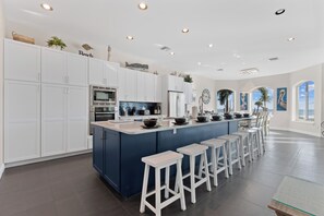 Une cuisine moderne avec des meubles blancs, un îlot central doté de tabourets et une vue sur l’océan grâce à de grandes fenêtres.