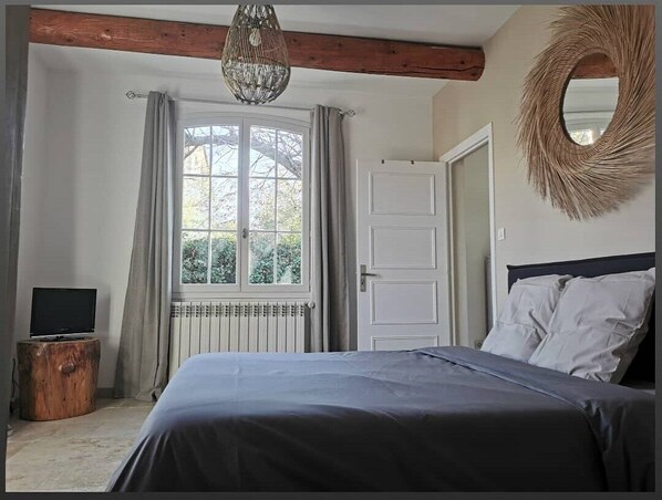 A bedroom with a bed, a television on a wooden stool, a window with curtains, a door, and a woven wall hanging.