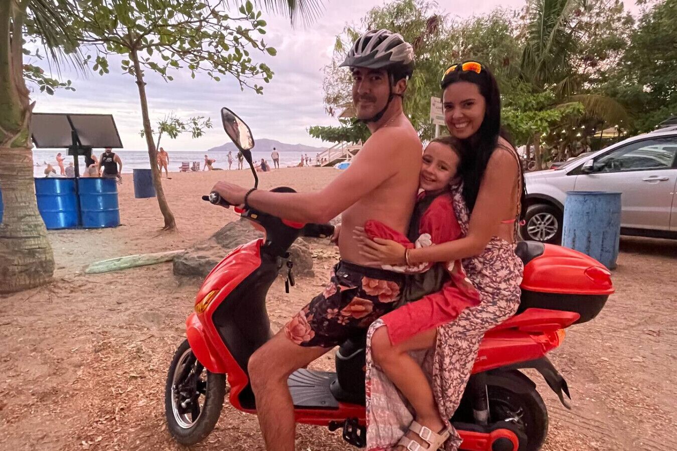 Une famille sur un scooter rouge, sur une plage bordée de palmiers, avec une voiture garée à proximité.