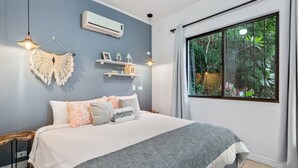 A bedroom with a bed, bedside tables, a hanging macrame wall decoration, a shelf with decorative items, and a window with a view of greenery. - Stylish + Fast Wifi+ IPTV+ Fully Equipped (Tamarindo)