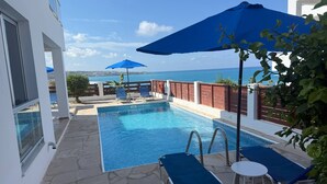A pool area with blue lounge chairs and umbrellas, a view of the sea, and a brick wall. - Villa Aqua Sunrise 1km From Beach With Sea Views (Coral Bay Peyia Paphos)