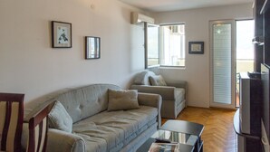 A living room with a sofa, armchair, and a coffee table. There are two windows with blinds, a door, and a TV on a stand. - Apartment Blue (Budva)