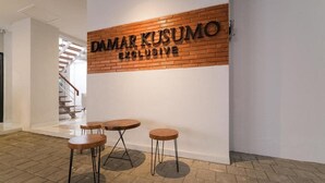 Un intérieur moderne avec une enseigne indiquant « DAMAR KUSUMO EXCLUSIVE » sur un mur de briques, un escalier en arrière-plan et trois tables rondes avec des tabourets.