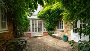 A stone patio with a metal table and chairs, surrounded by lush greenery and a brick wall.