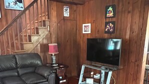 A living room with a wooden staircase, a black leather sofa, a TV on a stand, and a patterned rug.