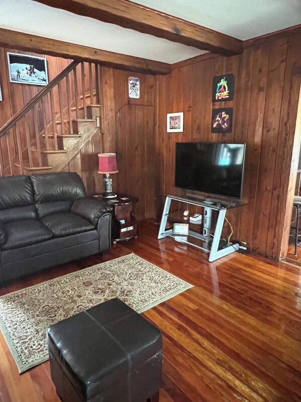 A living room with a wooden staircase, a black leather sofa, a TV on a stand, and a patterned rug.