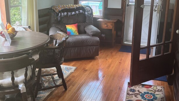 A living room with a wooden floor, a sofa, a dining table with chairs, and a door leading outside.