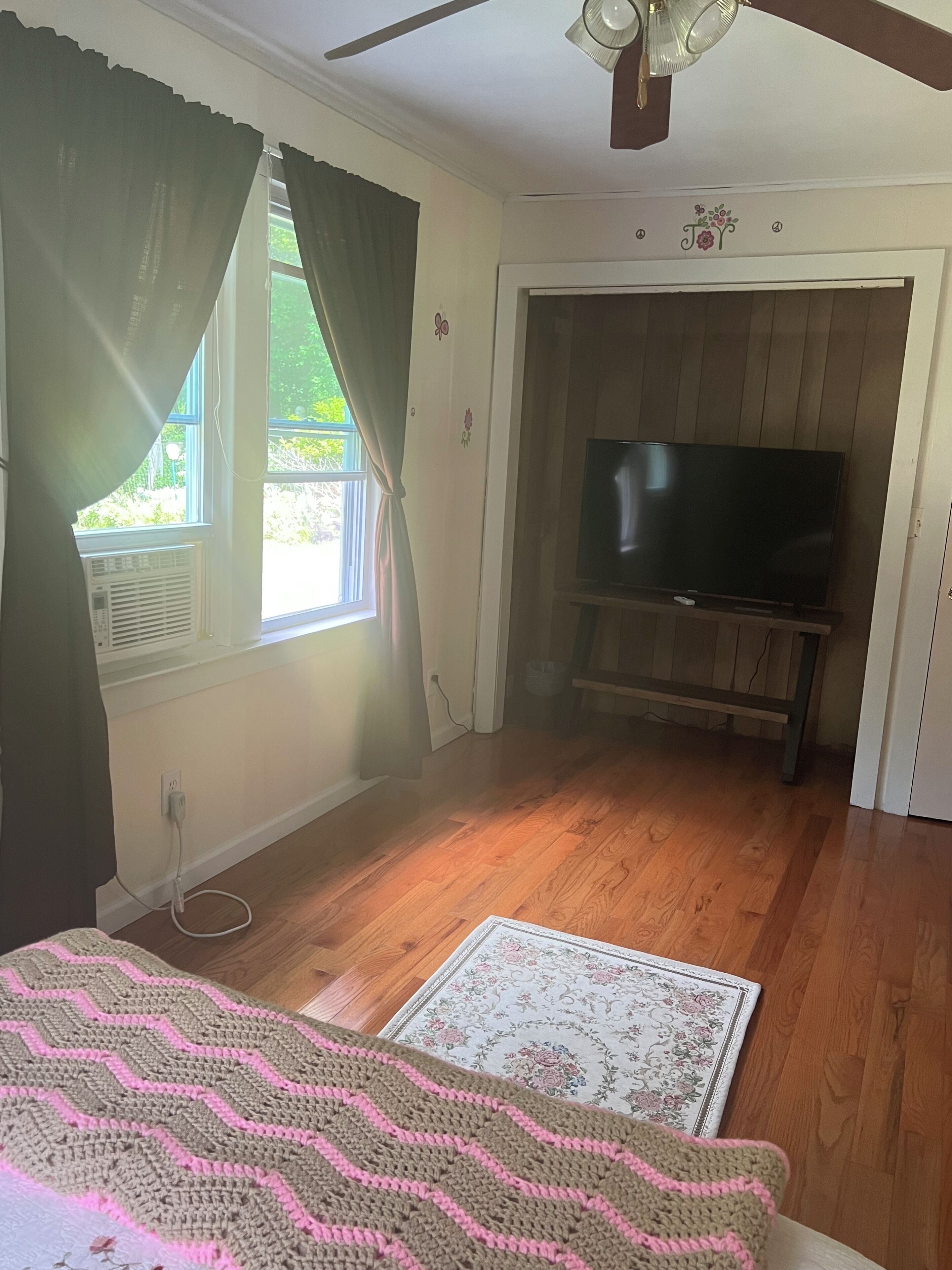 Ein Zimmer mit Holzboden, einem Fernsehgerät auf einem Schrank, einem Fenster mit Vorhängen und einer Tür mit floralen Verzierungen.