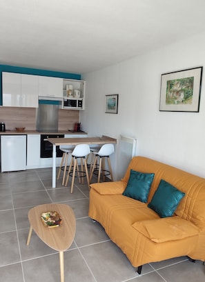 A modern kitchen with white cabinets, a stainless steel refrigerator, and a dining area with a wooden table and four white chairs. There is a mustard-colored sofa with teal cushions and a small wooden stool with a bowl of fruit.