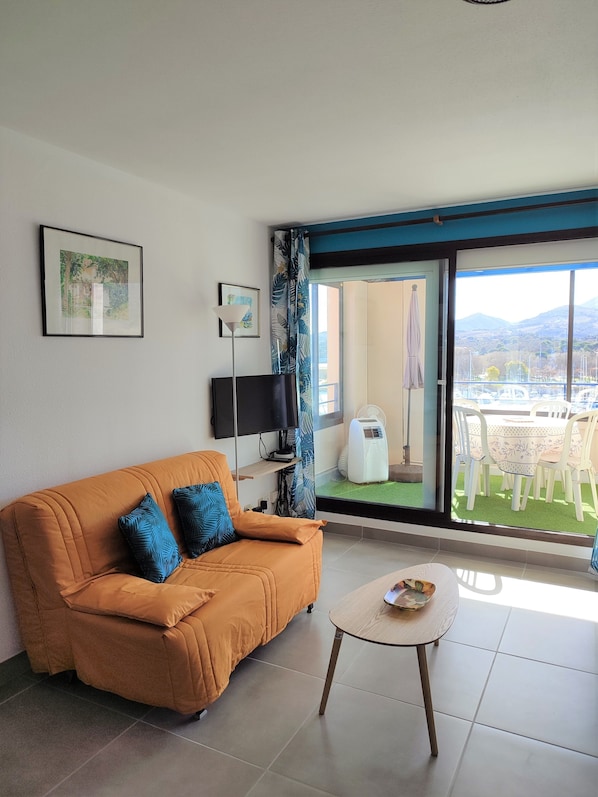 A living room with a sofa, a small table, a TV, and a view of a balcony with outdoor furniture.