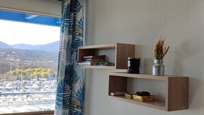 Ett rum med utsikt över en småbåtshamn, ett fönster med blå gardiner och trähyllor med diverse föremål.