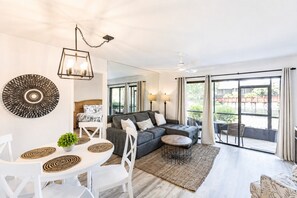 A modern living room with a sofa, dining table, and chairs, a ceiling fan, and large windows.