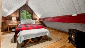 A cozy bedroom with a sloped ceiling, a bed with a red and black checkered blanket, two bedside tables with lamps, and a wood-burning stove.