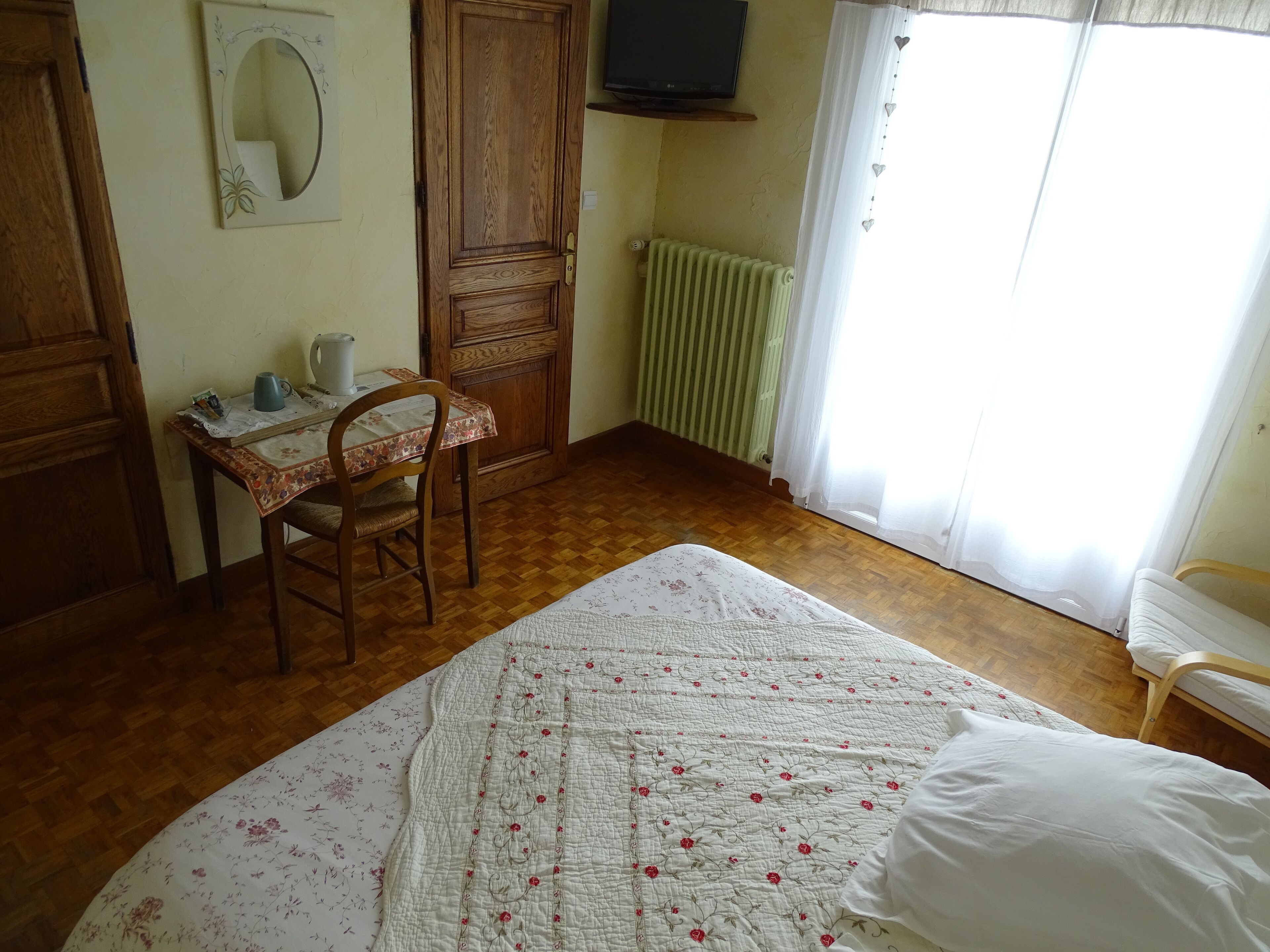 A bedroom with a wooden floor, a bed with a floral quilt, a wooden desk with a chair, a radiator, and a window with curtains.