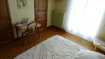 A bedroom with a wooden floor, a bed with a floral quilt, a wooden desk with a chair, a radiator, and a window with curtains.