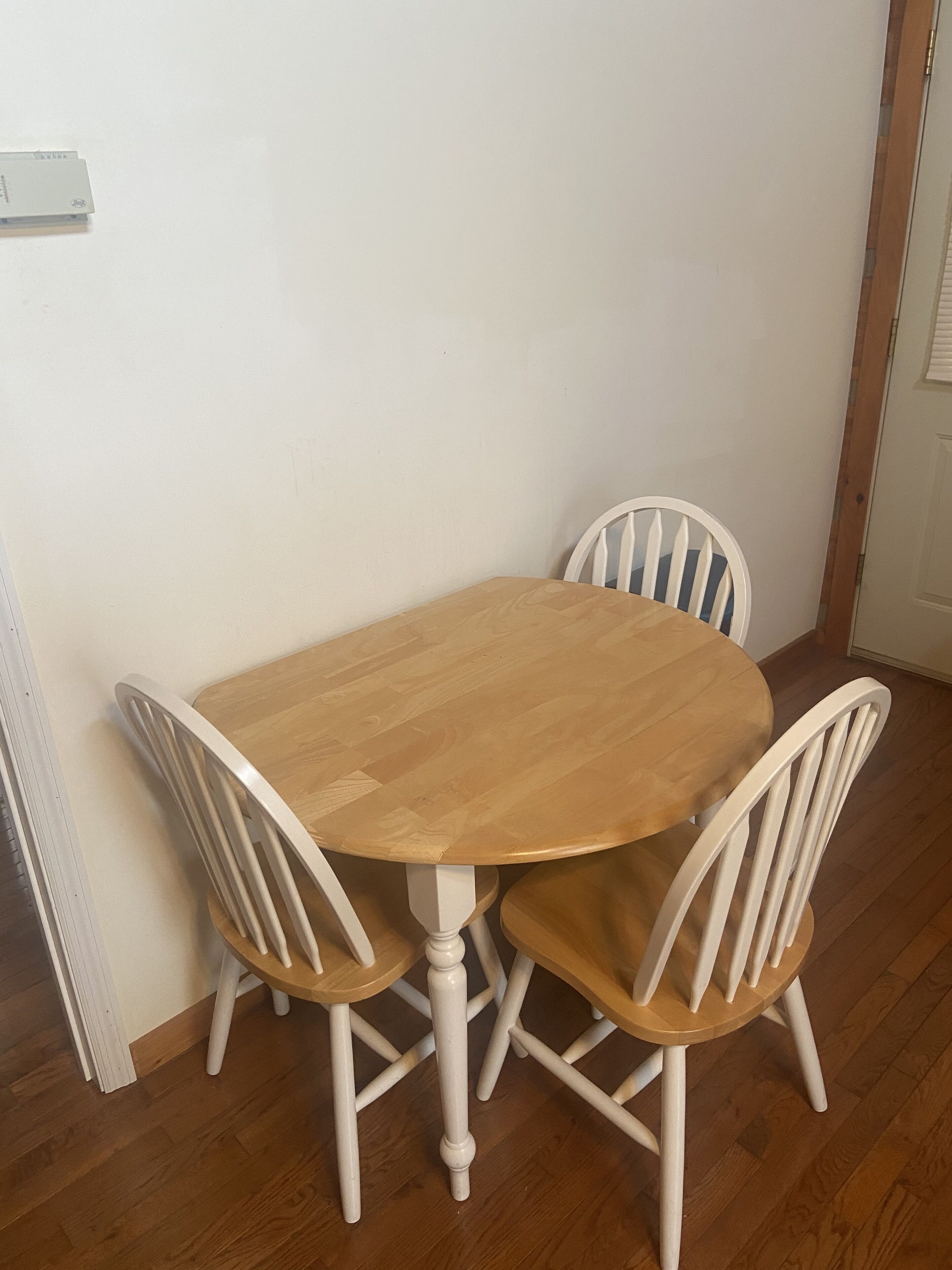 A dining table with four white chairs, a wooden floor, and a white wall.