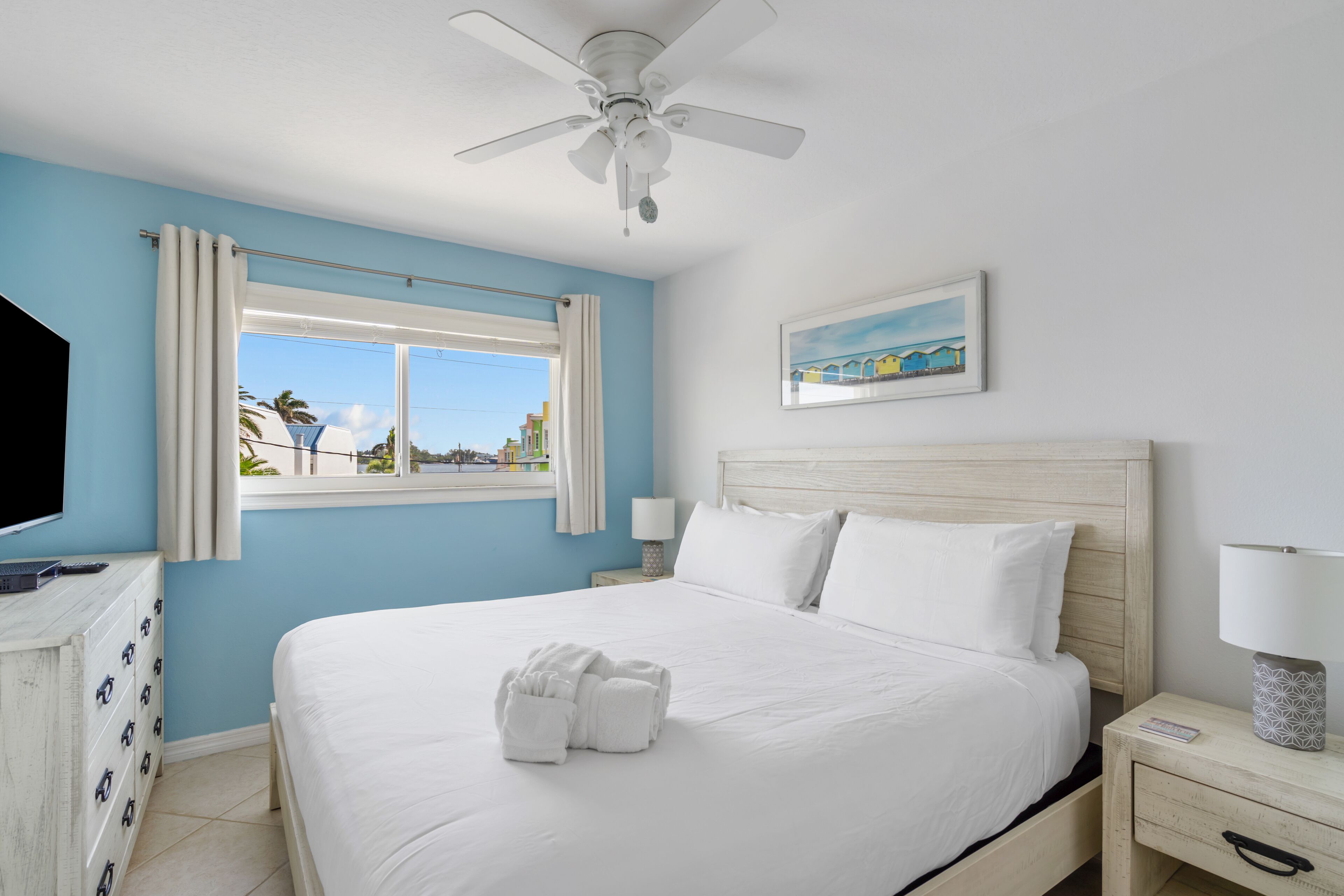 A bedroom with a bed, a ceiling fan, a window with curtains, a bedside table with a lamp, and a painting on the wall.