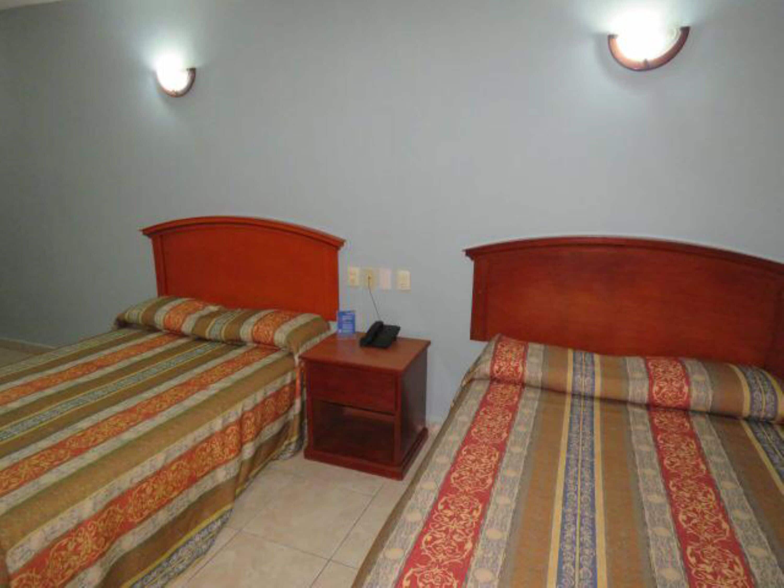 Une chambre avec deux lits, chacun recouvert d’une couvre-lit à rayures et doté d’une tête de lit en bois. Une table de chevet avec un téléphone et une lampe se trouve entre les deux lits.