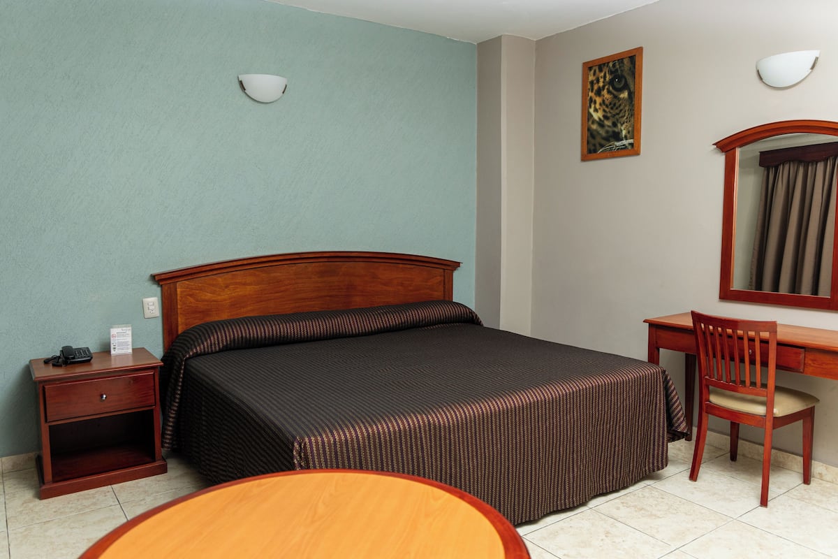 Una habitación de hotel con cama, escritorio, silla, mesita de noche y lámpara de pared.