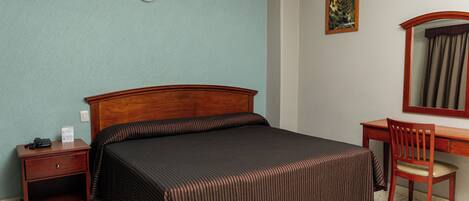 Quarto de hotel com cama, escrivaninha, cadeira, mesa de cabeceira e luminária na parede.