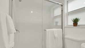 Un baño moderno con cabina de ducha de vidrio, azulejos blancos, un inodoro y una pequeña planta en maceta sobre una estante.