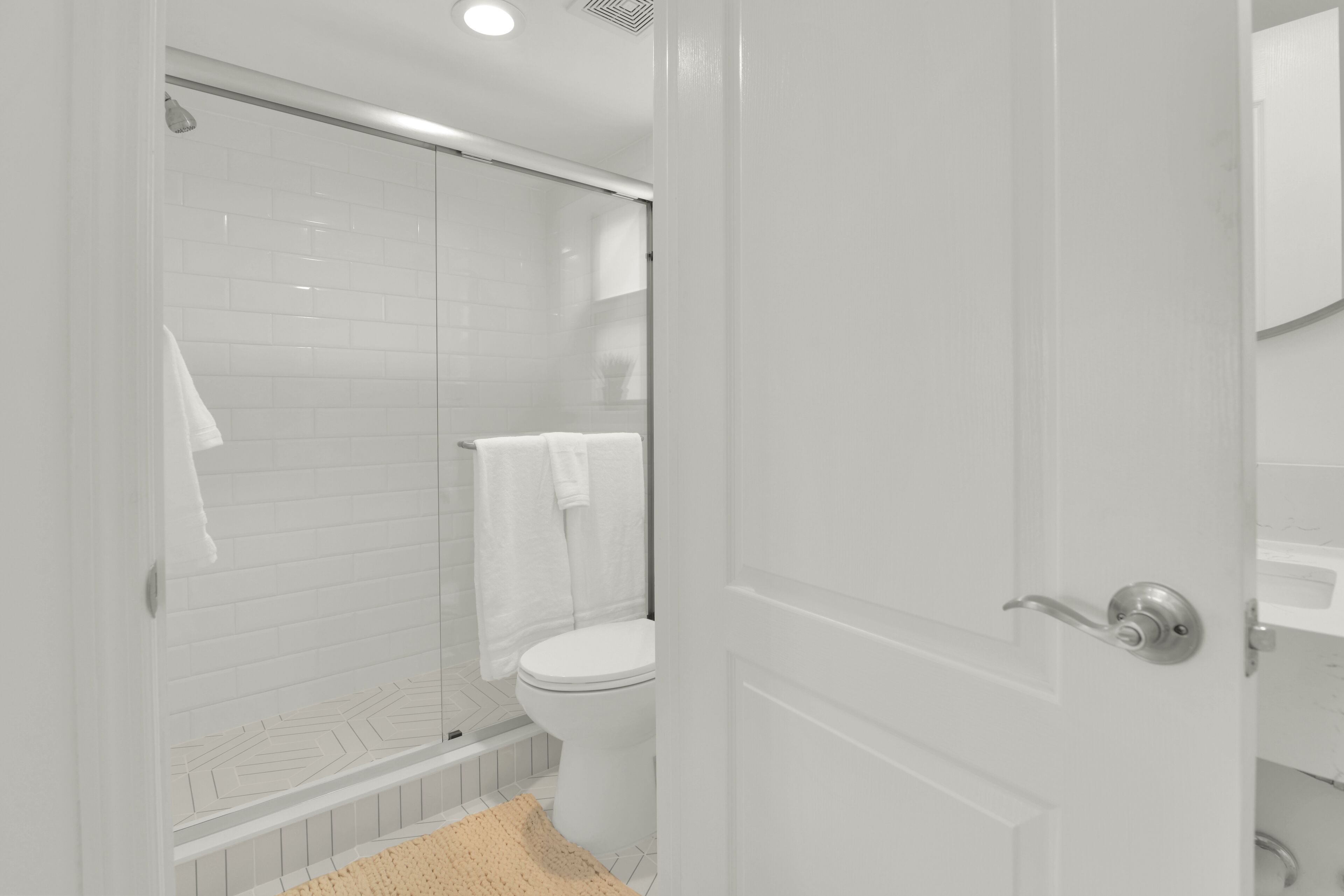 A bathroom with a glass shower enclosure, a white toilet, and a beige bath mat.