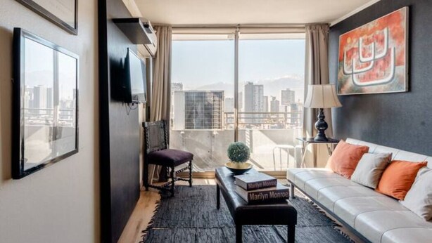 A modern living room with a sofa, a coffee table, a TV, and a view of the cityscape through large windows. - A2 lindo y comodo 2D 2 B full equipado hasta 6 pax metro en la puerta (Santiago)