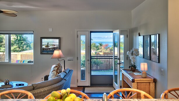 A living room with a sofa, dining table, and chairs, a balcony with a view of the ocean, and a door leading outside. - Kimo's Getaway (Kailua-Kona)