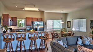 A modern kitchen with wooden cabinets, a stainless steel refrigerator, and a bar area with stools. There is a dining table with chairs and a sofa in the living area. - Kimo's Getaway (Kailua-Kona)