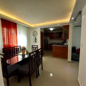 A modern dining area with a wooden table and chairs, a wall-mounted clock, and a kitchen in the background.