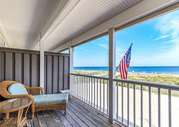 A wooden deck with wicker furniture and an American flag, overlooking a beach.