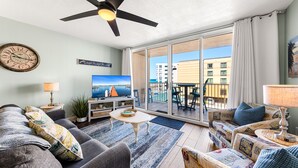 Ein Wohnzimmer mit einer Couch, Sesseln, einem Couchtisch, einem Fernseher und einem Balkon mit Blick auf Gebäude.