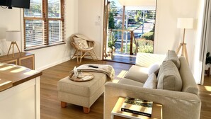 A modern living room with a sofa, ottoman, and wicker chair. There is a television mounted on the wall, a floor lamp, and a view of the outside through a glass door. - 1 Hour from Sydney 500 meters to Terrigal beach, restaurants and night life. (Terrigal)
