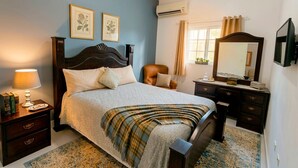 Una camera da letto con un letto, comodino, lampada, cassettiera, specchio e stampe botaniche incorniciate alle pareti.