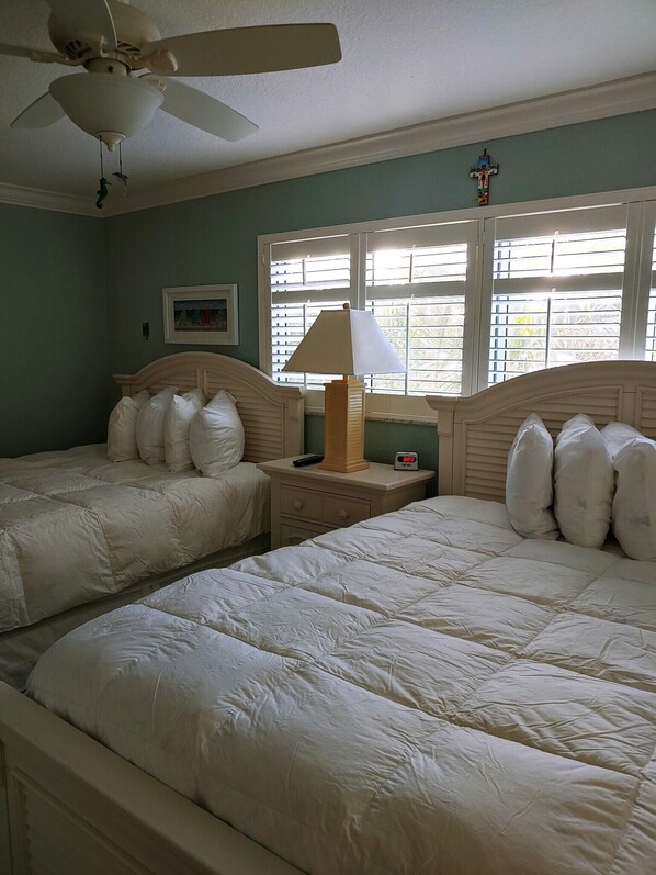 A room with two beds, a ceiling fan, a window with blinds, a bedside table with a lamp, and a clock.