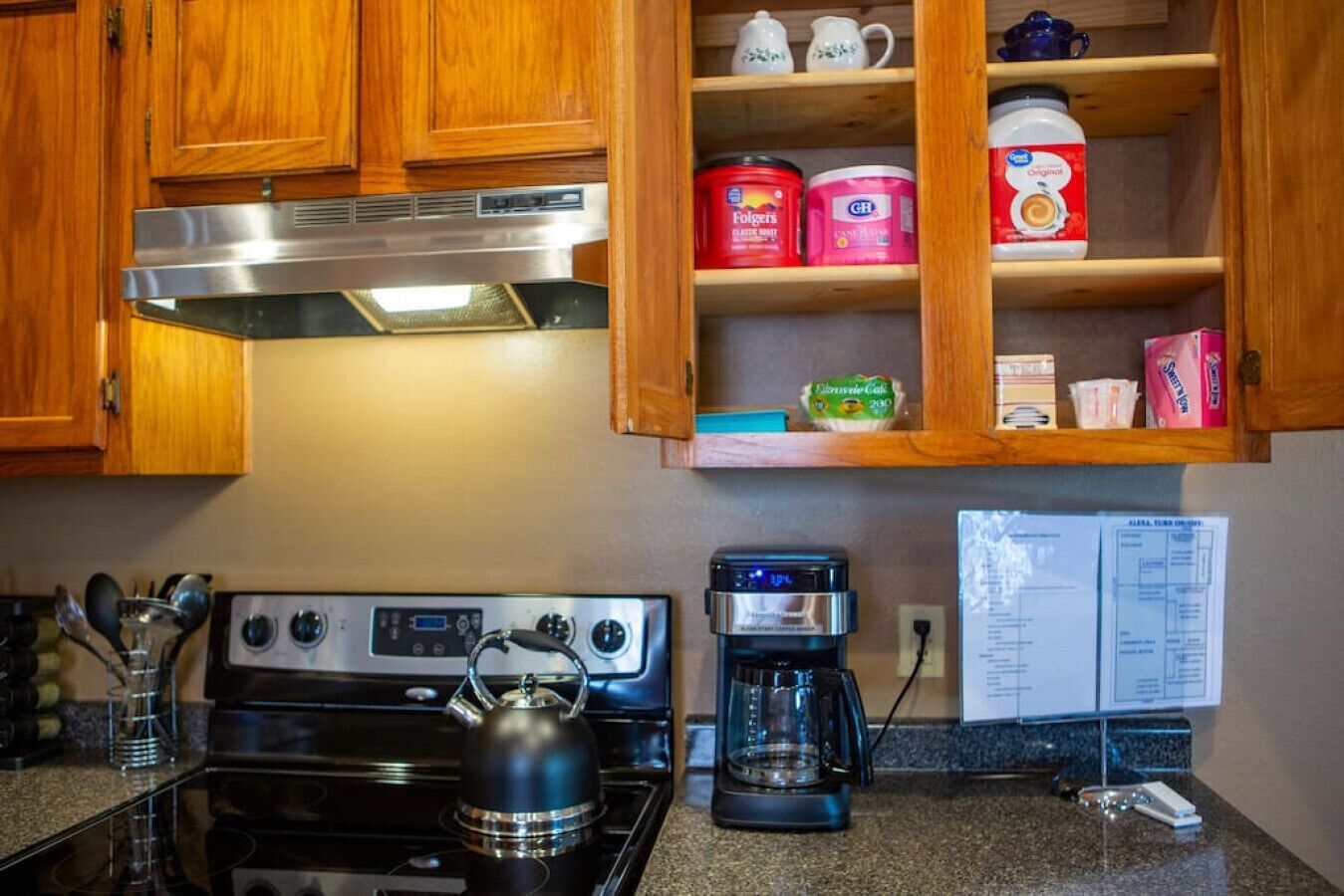 A kitchen with wooden cabinets, a stove, a kettle, a coffee maker, and various food items on shelves.